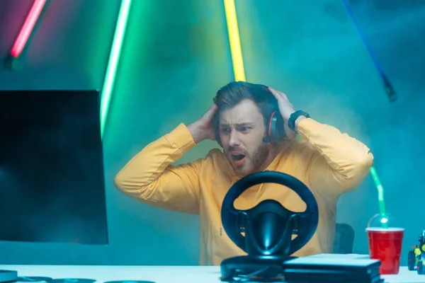 Shocked and handsome man in headphones playing video game with steering wheel — Stock Photo