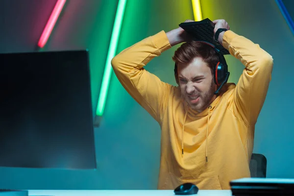 Angry and handsome man in headphones holding computer keyboard and looking at computer monitor — Stock Photo