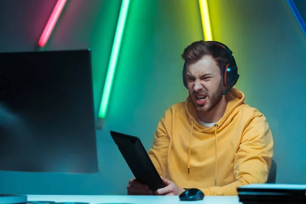 Angry and handsome man in headphones holding computer keyboard and looking at computer monitor — Stock Photo