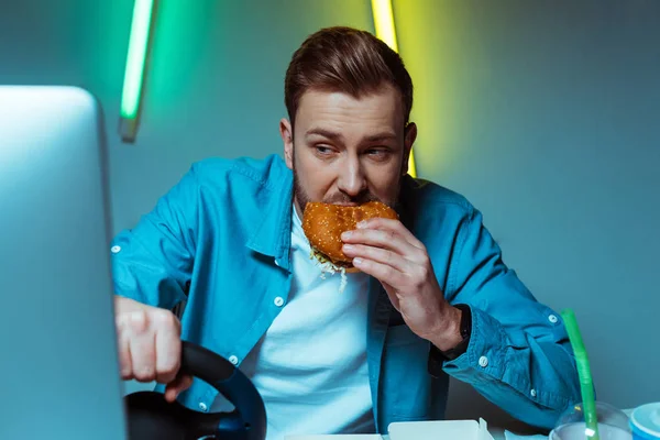 Hombre guapo jugando videojuego con volante y comer hamburguesa - foto de stock