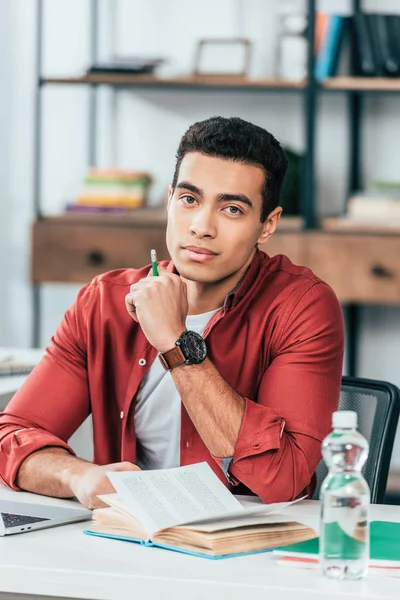 Hermosa estudiante morena con libro sentado en la mesa y sosteniendo el lápiz - foto de stock