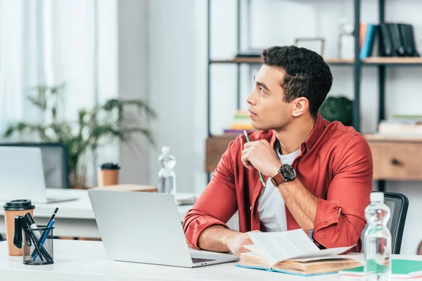 Étudiant pensif en chemise rouge regardant loin tout en utilisant un ordinateur portable — Photo de stock