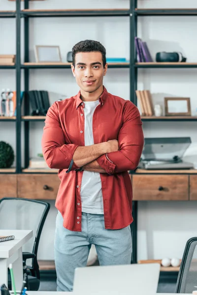 Joyful young man in red shirt standing with crossed arms at workplace — Stock Photo