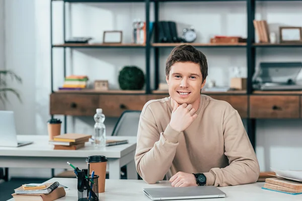 Estudante sorrindo sentado à mesa com laptop e olhando para a câmera — Fotografia de Stock