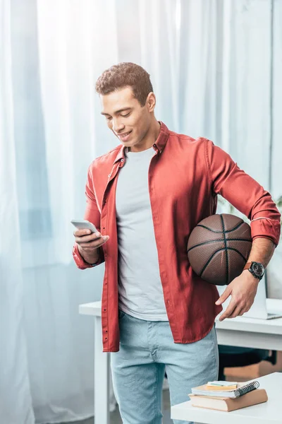 Брюнетка молодой человек в красной рубашке с баскетбольным мячом держа смартфон — стоковое фото