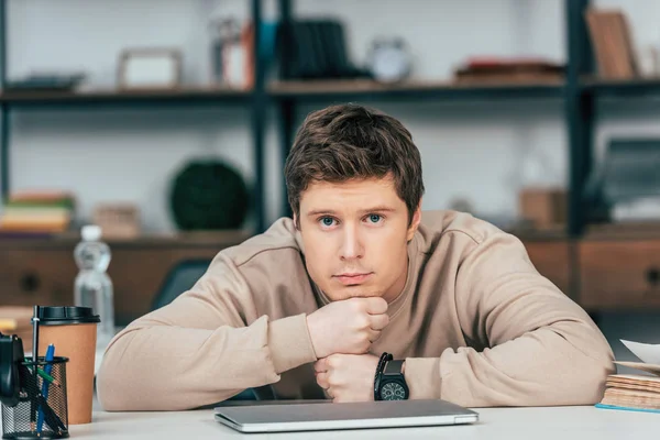 Estudiante molesto en reloj de pulsera sentado con los brazos cruzados cerca de la computadora portátil y mirando a la cámara - foto de stock