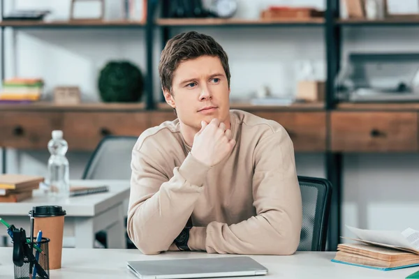 Étudiant pensif assis à table avec ordinateur portable et livre — Photo de stock