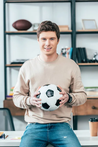 Jovem sorridente sentado na mesa e segurando bola de futebol no local de trabalho — Fotografia de Stock