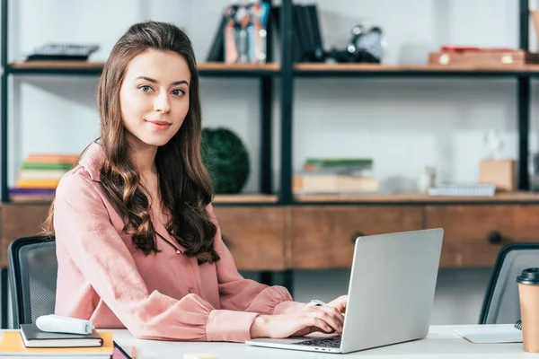 Fille attrayante en chemisier rose en utilisant un ordinateur portable et en regardant la caméra — Photo de stock