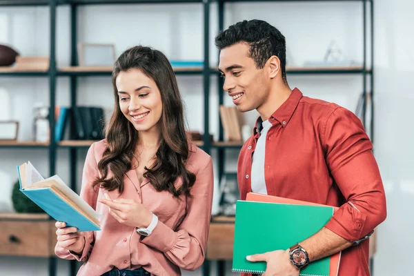 Dos estudiantes sonrientes multiétnicos con carpetas y libro preparándose para las lecciones - foto de stock