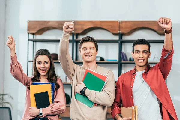 Drei fröhliche multiethnische Studenten mit Notizbüchern und Büchern in der Hand und mit erhobenen Fäusten — Stockfoto