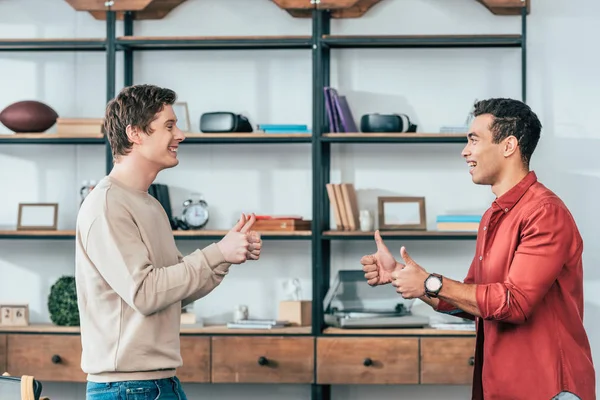 Seitenansicht von zwei multiethnisch lächelnden Freunden, die Daumen nach oben zeigen und einander anschauen — Stockfoto