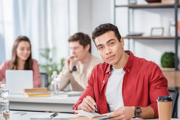 Cheerful student sitting at desk with book and looking at camera — Stock Photo