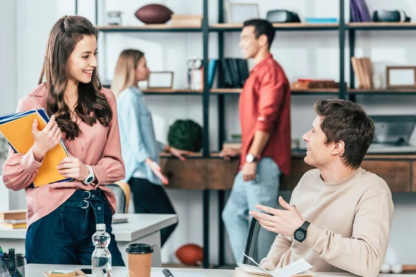 Muchacha atractiva sosteniendo cuadernos y hablando con un amigo con sonrisa - foto de stock