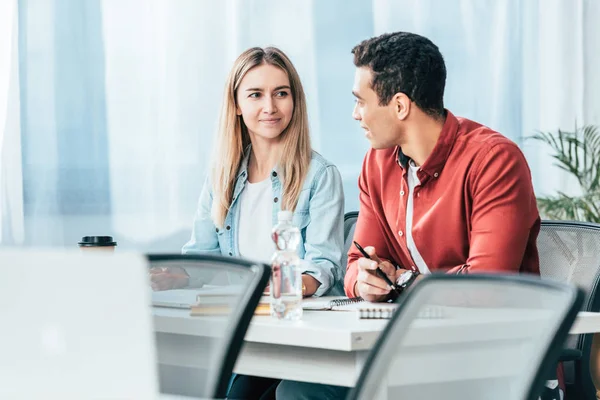 Multicultural students in casual clothes sitting at desk and looking at each other — Stock Photo