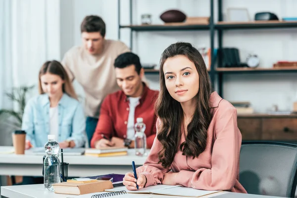 Pretty student writing in notebook and looking at camera — Stock Photo