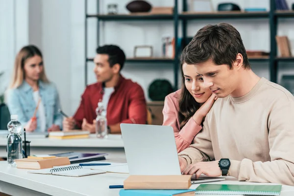 Estudiantes sonrientes sentados en escritorios y usando laptop mientras estudian en el aula - foto de stock