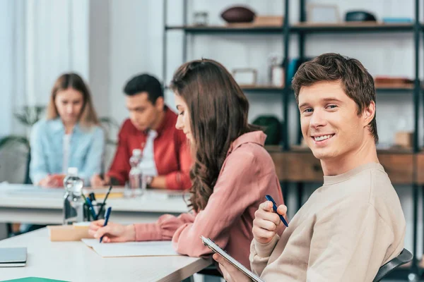 Estudiante feliz sosteniendo cuaderno con bolígrafo y mirando a la cámara con sonrisa - foto de stock