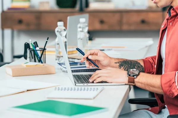 Vue recadrée de l'étudiant avec tatouage en chemise rouge à l'aide d'un ordinateur portable au bureau — Photo de stock