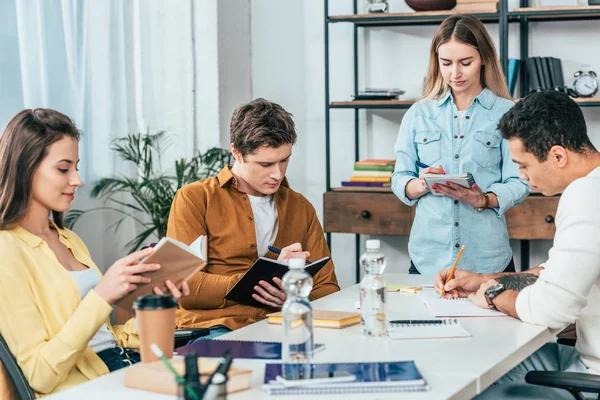 Quattro studenti multietnici che leggono libri e scrivono in quaderni a tavola — Foto stock