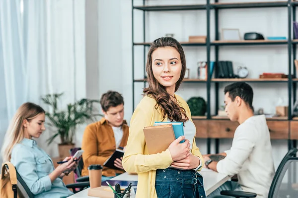 Encantador estudiante rizado de pie cerca de la mesa y sosteniendo libros - foto de stock