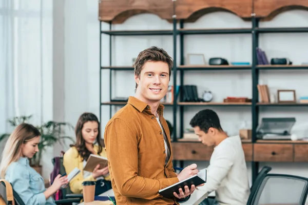 Lächelnder Student im Hemd blickt in die Kamera, während er in Notizbuch schreibt — Stockfoto