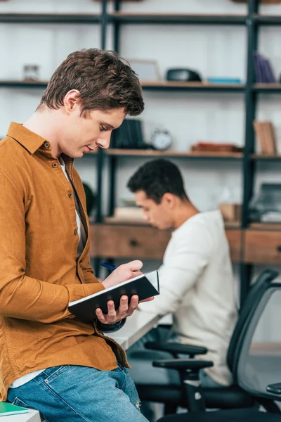 Konzentrierter Student in Hemd und Jeans, der in Notizbuch schreibt — Stockfoto