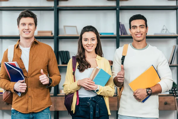 Drei multiethnisch glückliche Studenten, die Notizbücher halten und Daumen nach oben zeigen — Stockfoto