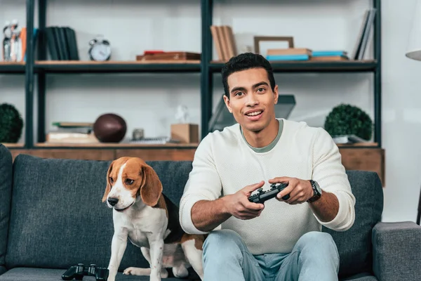 Junger Mann mit Hund sitzt auf Sofa und hält Steuerknüppel — Stockfoto