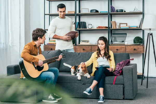 Tres amigos multiculturales con perro beagle tocando la guitarra en la sala de estar - foto de stock