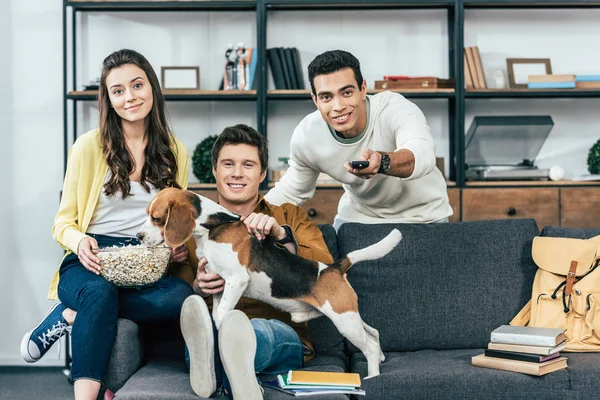 Три мультиэтнических улыбающихся друга с собакой и попкорном, сидящих на диване и смотрящих телевизор — стоковое фото