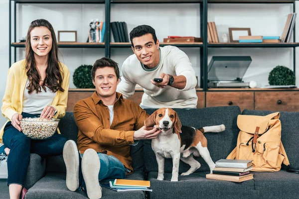Three smiling multicultural friends with dog and popcorn sitting on sofa and watching tv — Stock Photo