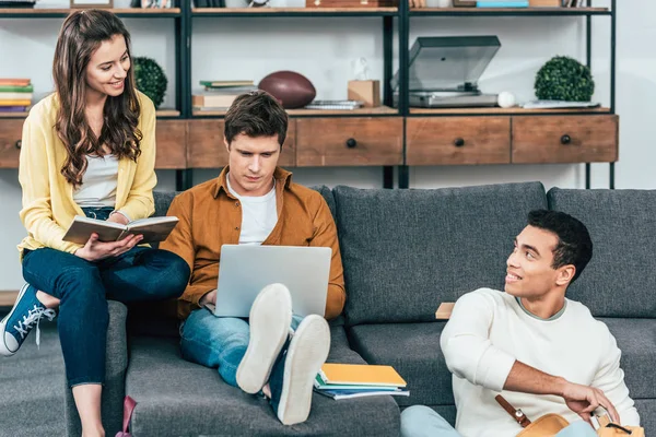 Tres estudiantes multiétnicos con cuadernos y portátil estudiando mientras están sentados en el sofá - foto de stock