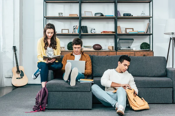 Tres estudiantes multiculturales con cuadernos y portátil estudiando mientras están sentados en el sofá - foto de stock