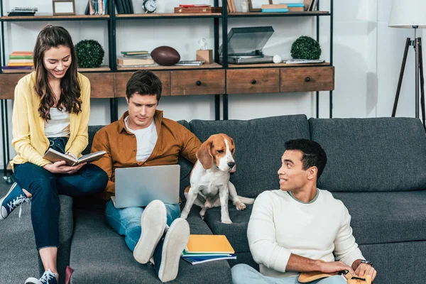 Tres estudiantes multiculturales con cuadernos y portátil estudiando mientras están sentados en el sofá - foto de stock