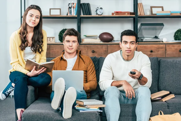 Três estudantes multiculturais com cadernos e laptop estudando e assistindo tv — Fotografia de Stock