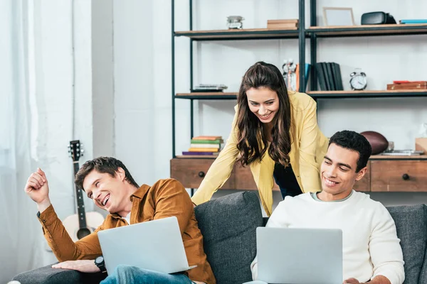 Three multicultural cheerful students sitting on sofa and uisng laptops at home — Stock Photo