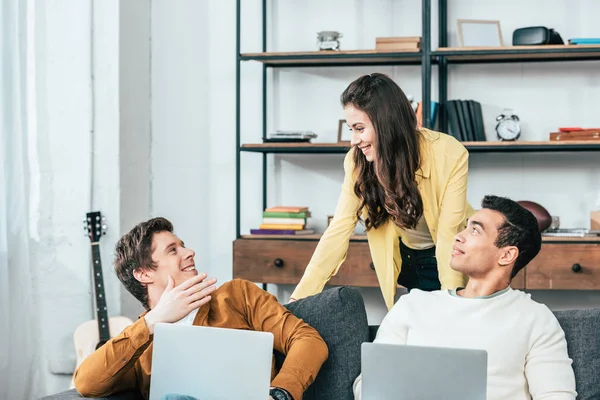 Three cheerful multiethnic students sitting on sofa and uisng laptops at home — Stock Photo