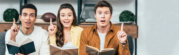 Panoramic shot of three multiethnic students with books pointing with fingers up in living room — Stock Photo