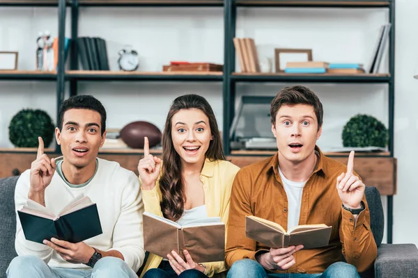 Drei multiethnische Studenten mit Büchern, die mit erhobenen Fingern im Wohnzimmer stehen — Stockfoto