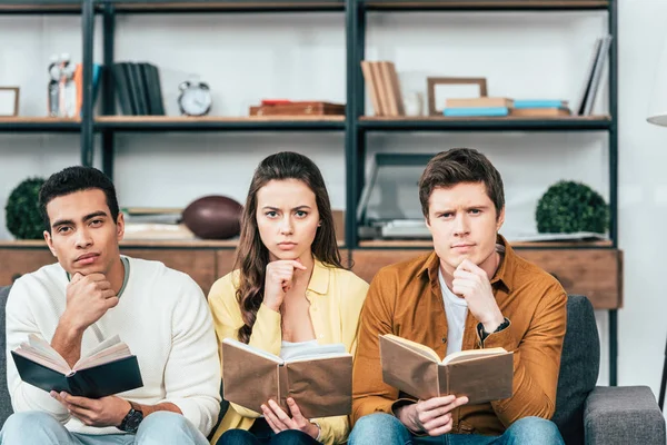 Tres estudiantes multiculturales sentados en el sofá y leyendo libros en el salón - foto de stock