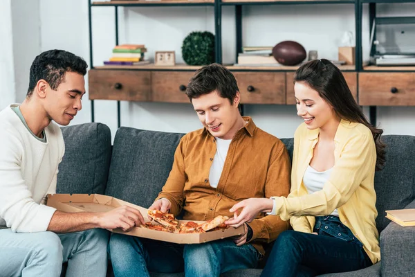 Tres alegres amigos multiétnicos sentados en el sofá y comiendo pizza juntos - foto de stock