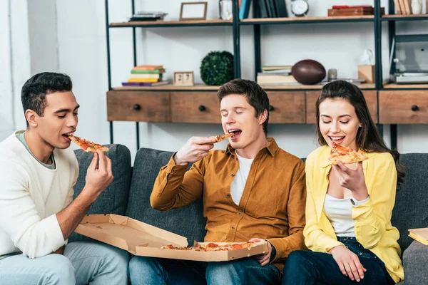 Three multiethnic joyful friends sitting on sofa and eating pizza together — Stock Photo