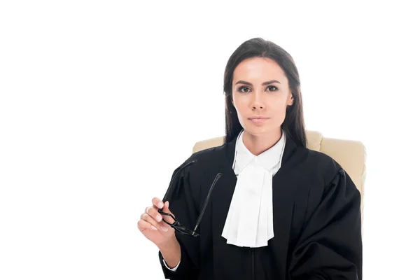 Hermoso juez en bata judicial sentado en sillón y sosteniendo gafas aisladas en blanco - foto de stock