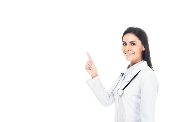 Medico sorridente in mantello bianco con stetoscopio puntato con dito isolato su bianco — Foto stock