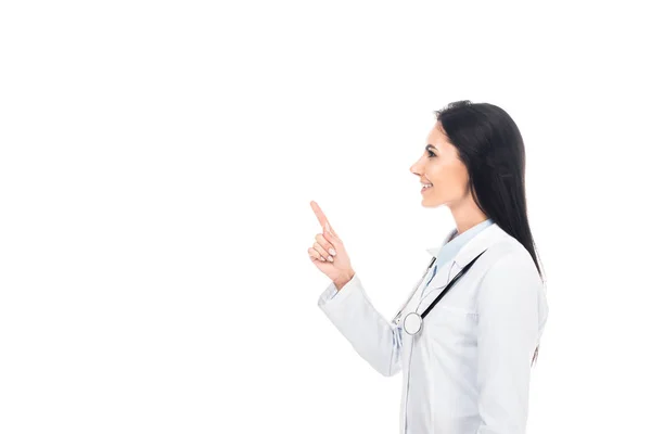 Вид сбоку улыбающегося врача в белом халате со стетоскопом, указывающим пальцем изолированным на белом — стоковое фото