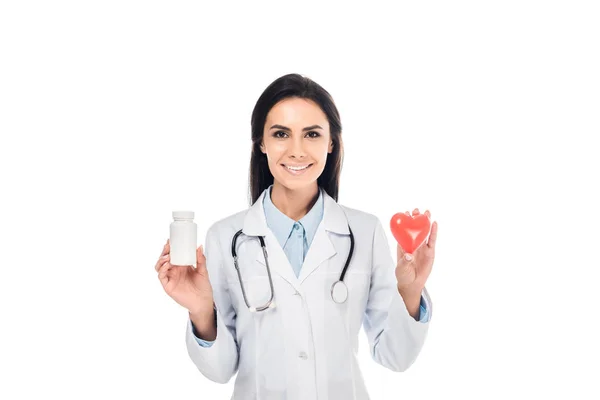 Doctor en bata blanca con estetoscopio sosteniendo píldoras y corazón de plástico aislado en blanco - foto de stock