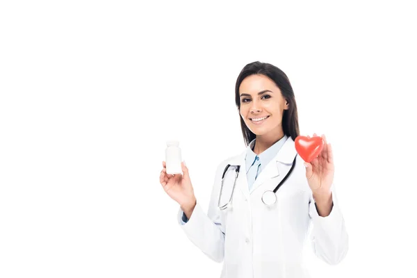 Доктор у білому пальто зі стетоскопом, що тримає таблетки та пластичне серце ізольовано на білому — стокове фото