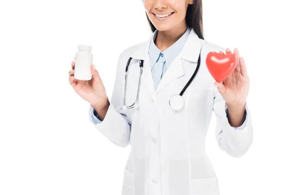 Vista recortada del médico en bata blanca con estetoscopio que sostiene las píldoras y el corazón plástico aislado en blanco - foto de stock