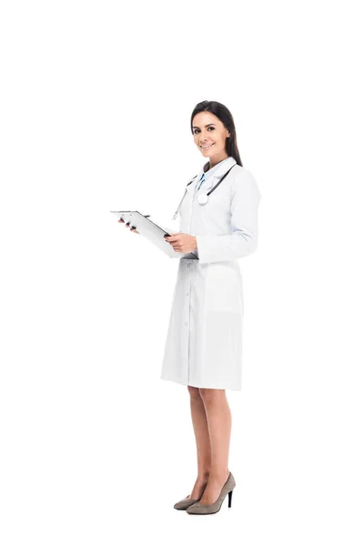 Ganzkörperansicht eines lächelnden Arztes in weißem Mantel, der Klemmbrett isoliert auf weiß hält — Stockfoto